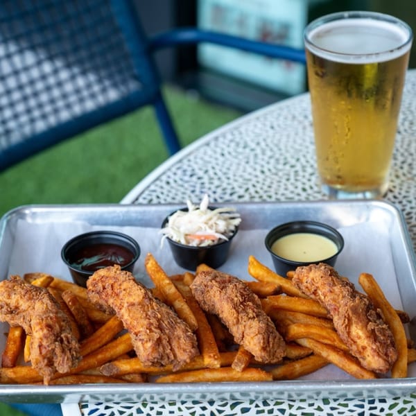 Chicken tenders and fries with coleslaw and dipping sauces on a metal tray with a beer.