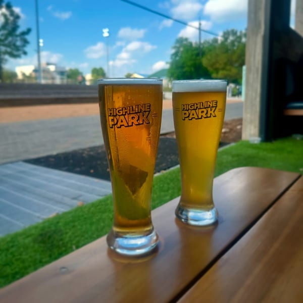 Two Highline Park branded beer glasses filled with golden beer on an outdoor wooden table.