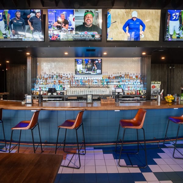 A long bar with brown stools, a tiled blue and white floor, and multiple TVs showing sports.