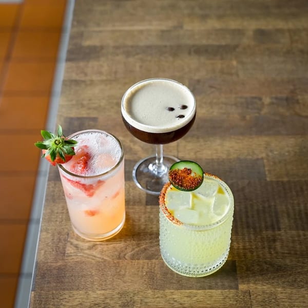 Three vibrant cocktails on a wooden table: a strawberry drink, an espresso martini, and a spicy margarita.
