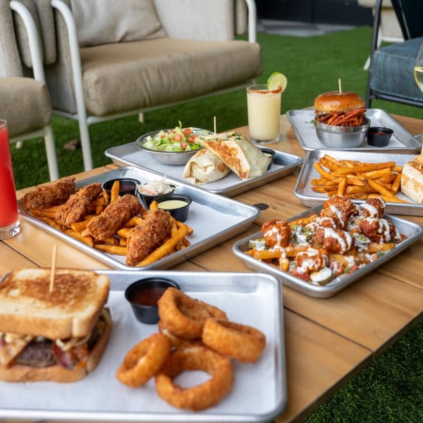 A wooden table laden with various bar-and-grill dishes and drinks on a patio with green turf.