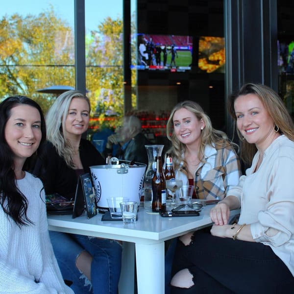 Four smiling women seated at an outdoor table with drinks and a bucket of seltzers at Highline Park.