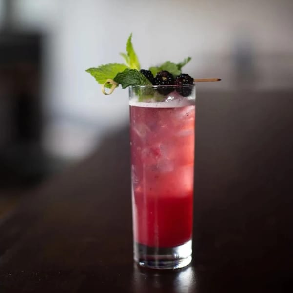 A tall glass of Highline Park's red cocktail, garnished with mint and blackberries, on a dark table.
