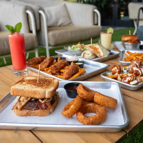 Highline Park's bar-&-grill dishes, including a burger, onion rings, chicken tenders, and drinks on an outdoor table.