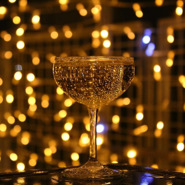 A coupe glass filled with a bubbly drink, set against a background of warm, blurred lights.