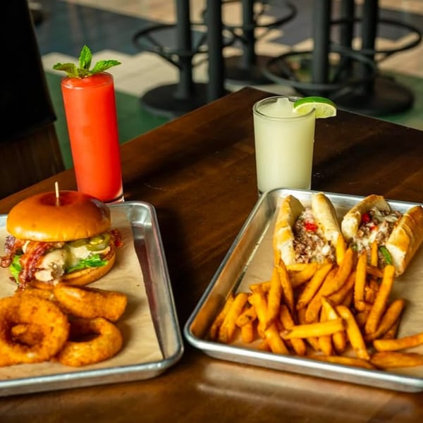 Highline Park's burger with onion rings, cheesesteak with fries, and two cocktails on a wooden table.