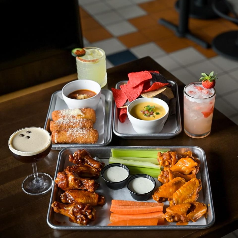 A table spread of Highline Park's bar-and-grill appetizers and cocktails, including wings, mozzarella sticks, and queso.