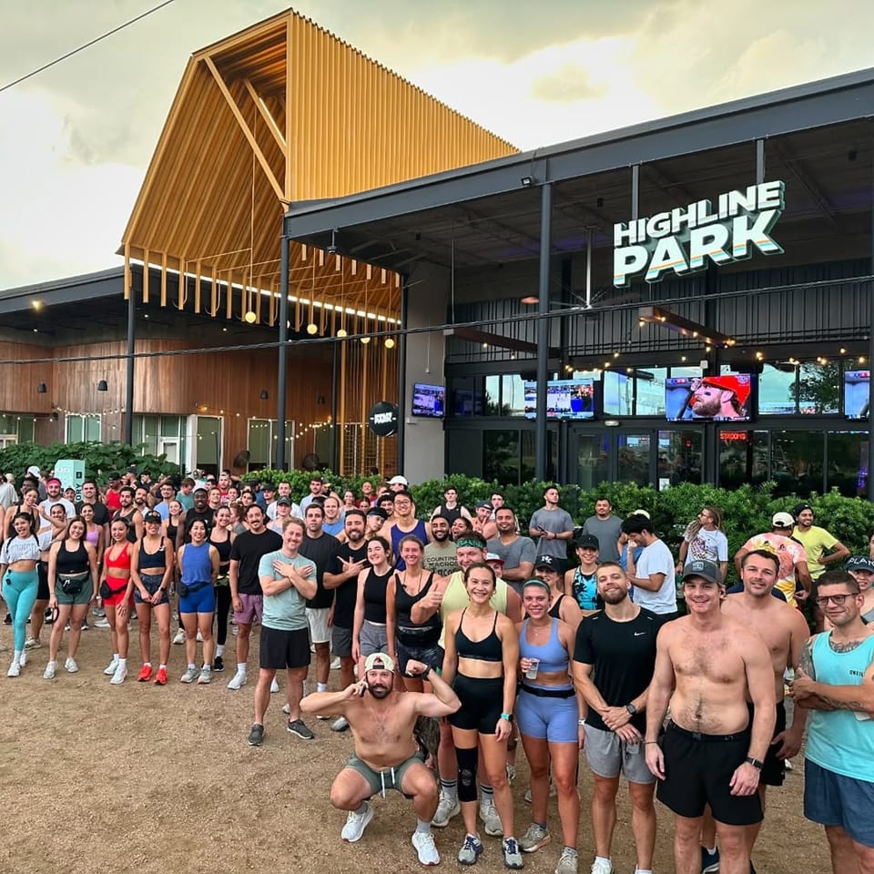 A large group of people gathered in front of Highline Park bar-&-grill, with a modern building and outdoor screens.