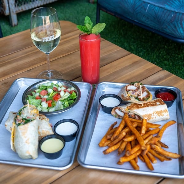Two trays of wraps, fries, and salad with drinks on a wooden table at Highline Park.