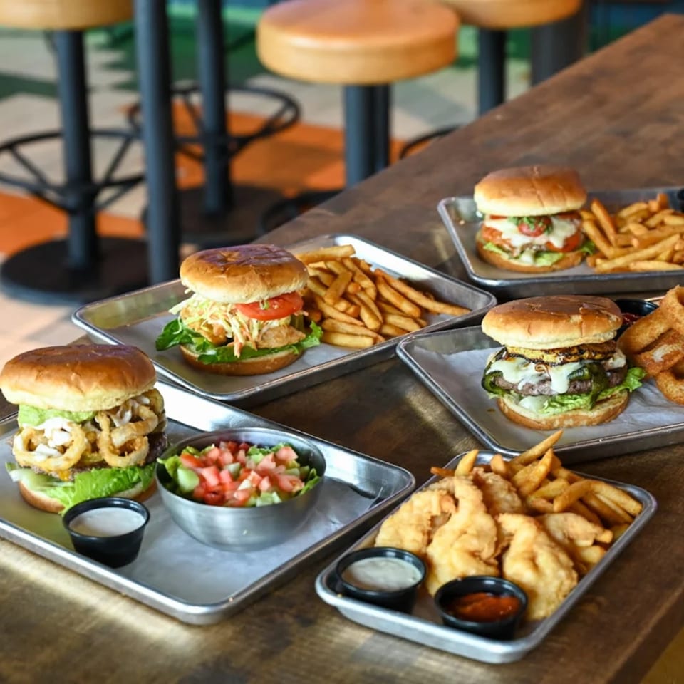 Several Highline Park bar-&-grill burgers, fries, and sides on metal trays on a wooden table.