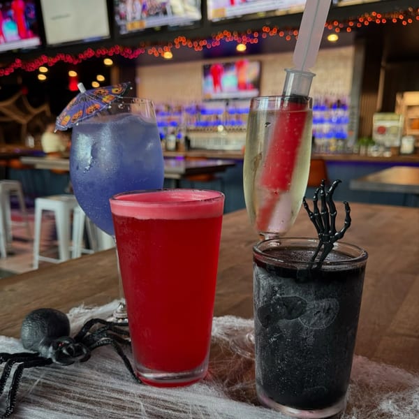 Four Halloween-themed drinks on a bar, with a spider decoration and string lights in the background.